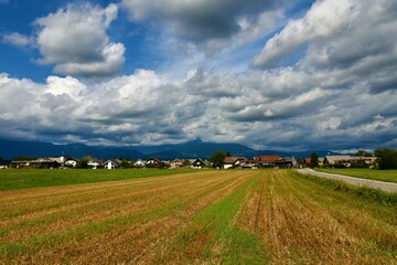 Harvested field next to a road and a village and mountains covered in clouds behind at Sorsko polje in Gorenjska, Slovenia