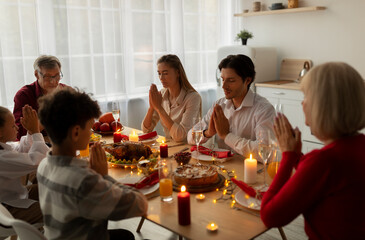 Extended family praying before Christmas holiday dinner, giving thanks to God at table indoors