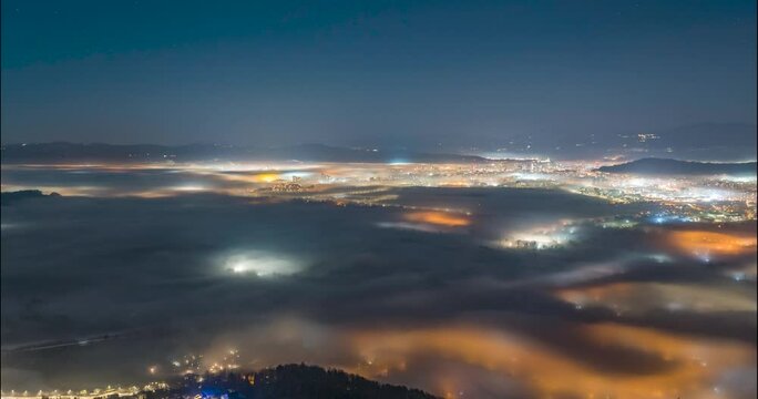 Time lapse of inversion low clouds or fog moving in Ljubljana basin at night. Big city under thin layer of mist in Slovenia. Traffic lights. Elevated or aerial view. Static shot