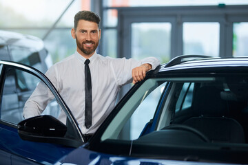 Fototapeta na wymiar Handsome bearded man in formal outfit visiting auto salon for choosing new vehicle. Happy male customer standing near open door of luxury car and smiling on camera. 