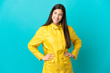 Teenager Brazilian girl wearing a rainproof coat over isolated blue background posing with arms at...