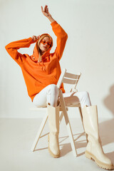 Full-length studio fashion portrait of playful confident woman wearing stylish orange hoodie, sunglasses, white skinny jeans, high boots, sitting, posing on chair.  - obrazy, fototapety, plakaty