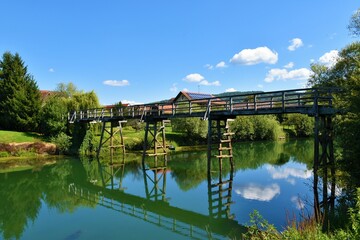 Fototapeta na wymiar Wooden bridge leading across Krka river at Kostanjevica na Krki in Dolenjska, Slovenia with a refelction of the sky and trees in water