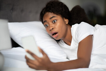 Shocked black woman looking at cellphone, laying in bed