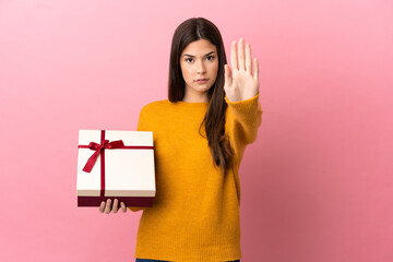 Teenager Brazilian girl holding a gift over isolated pink background making stop gesture