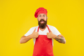 Surprised man in toque and apron giving double thumbs yellow background, cook