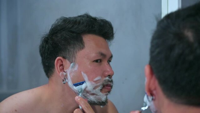 Adult man shaving with foam and manual razer.Mirror reflection young handsome smiling man touching beard, grooming in bathroom. Head shot close up happy asian man guy doing morning beauty routine.