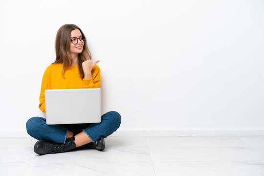 Young caucasian woman with a laptop sitting on the floor isolated on white background pointing to the side to present a product