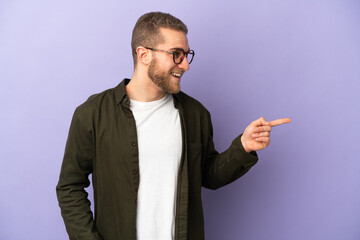 Young handsome caucasian man isolated on purple background pointing finger to the side and presenting a product