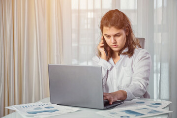 Disabled woman freelancer with cerebral palsy talks on mobile phone explaining project paper near laptop at table against window at home, sunlight.