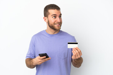 Young handsome caucasian man isolated on white background buying with the mobile with a credit card while thinking