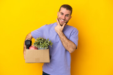 Young caucasian making a move while picking up a box full of things isolated on yellow background...