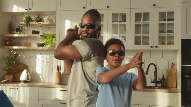 Cheerful good looking African father and adorable elementary age boy in sunglasses making flinger gun gesture, looking with friendly smiles, expressing carefree mood and happiness at home.