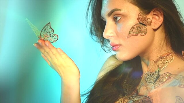 Beauty model girl in colorful bright neon lights posing, fashion make up with butterfly. Portrait of beautiful summer young woman in UV. Art design colorful make up with golden butterflies Slow motion