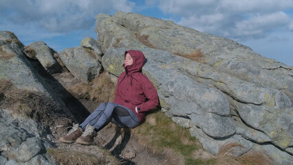 Exhausted tourist woman after hard trekking in mountains sits near large rock, relaxes in sun. Backpacker girl resting in mountains. Active traveler on background of gray rocks, blue sky with clouds