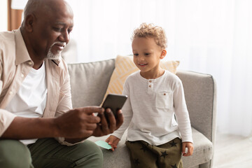 Happy Black Grandpa Showing Mobile Phone To Grandson At Home