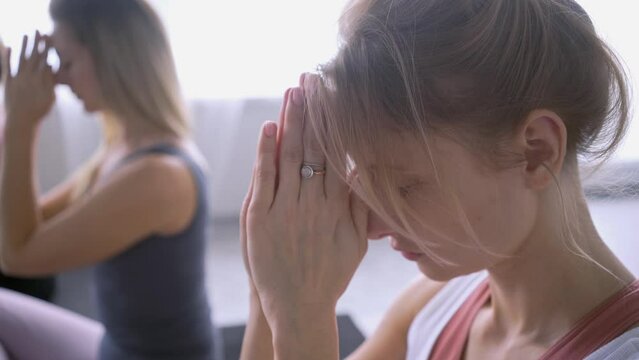 Close-up portrait concentrated woman showing praying gesture, holding palms before face with closed eyes, small female student group sitting on floor in light spacious gim