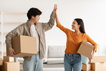 Young Asian couple with cardboard boxes high fiving each other in new home on moving day