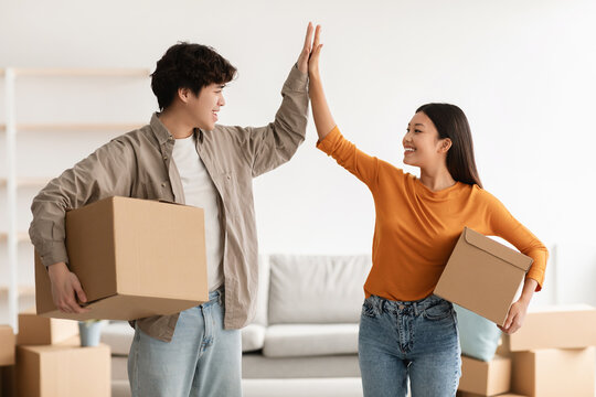 Young Asian couple with cardboard boxes high fiving each other in new home on moving day