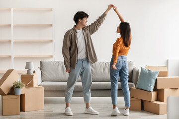 Loving millennial Asian couple dancing in their new home among carton boxes on moving day, copy space