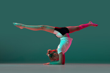 Handstand. Little flexible girl, rhythmic gymnastics artist training isolated on green studio background in neon pink light. Grace in motion, action. Doing exercises in flexibility.