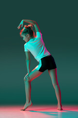 Little flexible girl, rhythmic gymnastics artist training isolated on green studio background in neon pink light. Grace in motion, action. Doing exercises in flexibility.