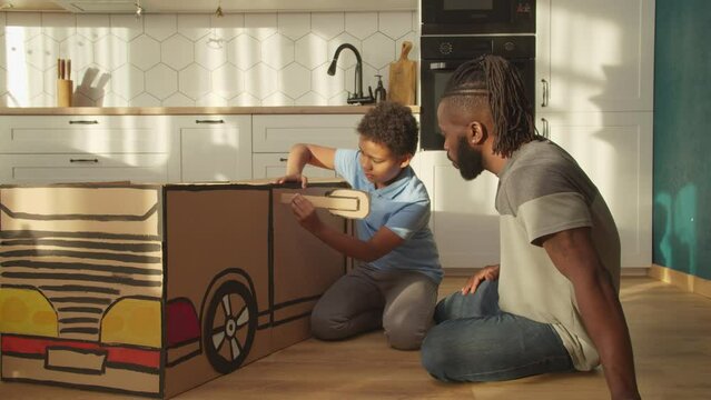 Affectionate stylish African father and adorable preadolescent boy sitting on floor, creating diy car toy from cardboard together and attaching toy car rear view mirror while enjoying leisure indoors.