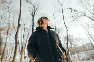 Fototapeta na wymiar Stylish lady in warm casual clothes stands in a snowy forest at sunset and looks away