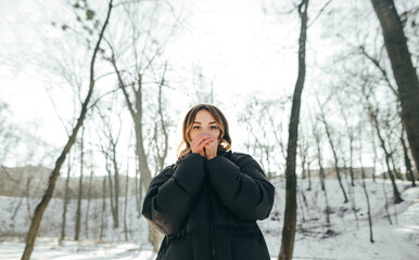 A frozen woman in a winter jacket stands in a snowy forest and breathes warm air in her palm to keep from freezing, looking into the camera against the backdrop of winter landscapes.