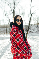 Winter portrait of stylish woman in casual clothes on the street in winter on a background of snow-covered park posing at camera in sunglasses.