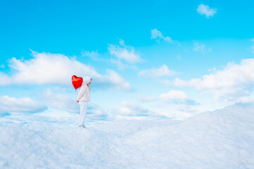 Portrait of young woman like angel looking down and red heart balloon walking on snow cover with...