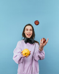 Positive girl in headphones juggles apples on a blue background with a smile on his face. Vertical