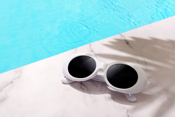 Vintage retro white sunglasses by the side of a pool with blue water with marble surface and palm...