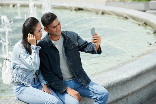 Smiling young couple sitting at fountain and taking selfie on smartphone