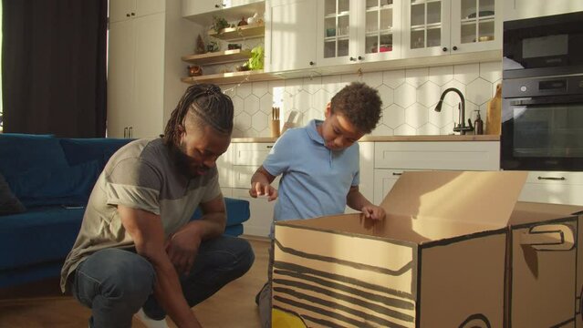 Busy cute preadolescent African American son and handsome father playing with big handcrafted toy cardboard car, having fun repairing broken vehicle with toy tools while relaxing at home.