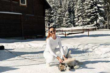 a beautiful girl in white clothes and sunglasses has fun playing with a fluffy and joyful husky on the background of nature in winter. dog on a leash.