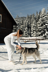 Naklejka premium a beautiful girl plays with a husky in the winter on the street against the background of the house and nature. fluffy husky plays with the mistress. girl with a dog in everyday life