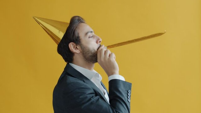 Portrait of happy Arab man wearing birthday hat blowing horn having fun alone on yellow color background. Holiday and people concept.