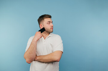 Adult man in a white T-shirt isolated on a blue background with a bank card in his hand, looks away and smiles.