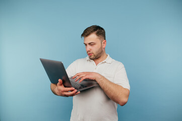 A man in a white T-shirt works on a laptop on a blue background with a serious face, looks at the screen and types text on the keyboard.