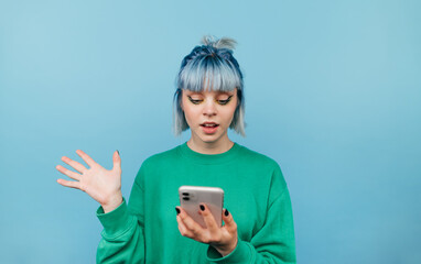 Surprised girl with blue hair uses a smartphone with a shocked face on a blue background and looks at the screen.