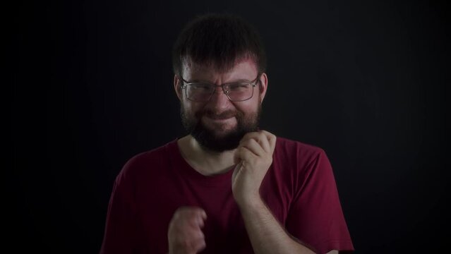 A bearded man wearing glasses and a burgundy T-shirt depicts the emotion of misunderstanding and spreads his arms to the sides against a black monochromatic background. Front view, slowmo.