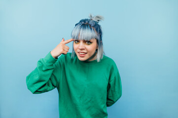 Funny girl in casual clothes and colorful hair shows her finger on her breasts and looks at the camera with an ironic look, isolated on a blue background.