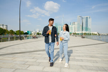 Happy excited young couple walking on river embarkment, drinking soft beverages, talking and joking around