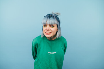 Cute girl with blue hair clenching her teeth looks into the camera and squints her eyes, isolated on a blue background, wearing a green sweatshirt.