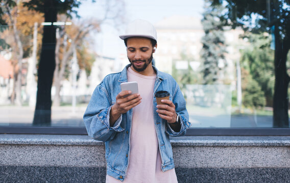 Hispanic hipster guy with caffeine beverage enjoying time for watching online video vlog on mobile website, male blogger with takeaway coffee to go connection to bluetooth on earbuds and smartphone
