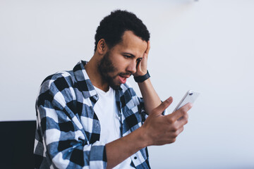 Emotional male employee receive email with bad news feeling shocked in office interior, unhappy hipster guy using smartphone for making online banking solving problems with internet connection