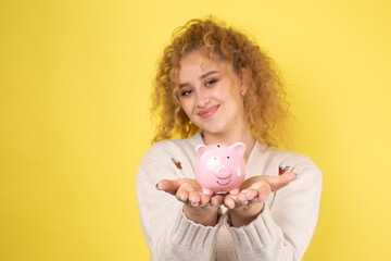 Fototapeta na wymiar A cute young girl with curly red hair holds a piggy bank, a pink piglet in her hands. The concept of wealth and safety of money.