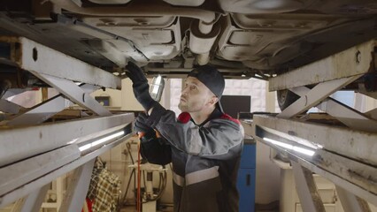 Mechanic walking under lifted car and inspecting car bottom with lamp while working in auto service garage