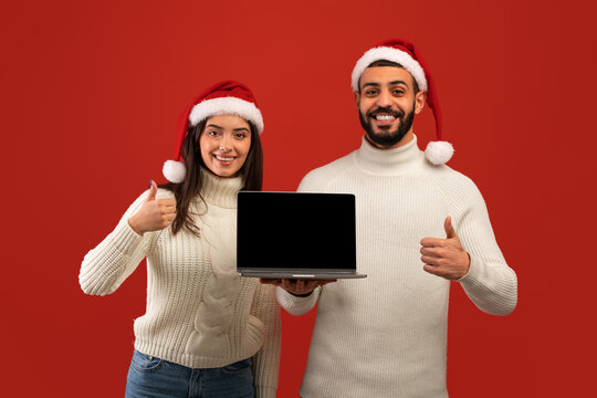 Mockup image of happy arab couple in Santa hats holding laptop with blank screen and showing thumbs up